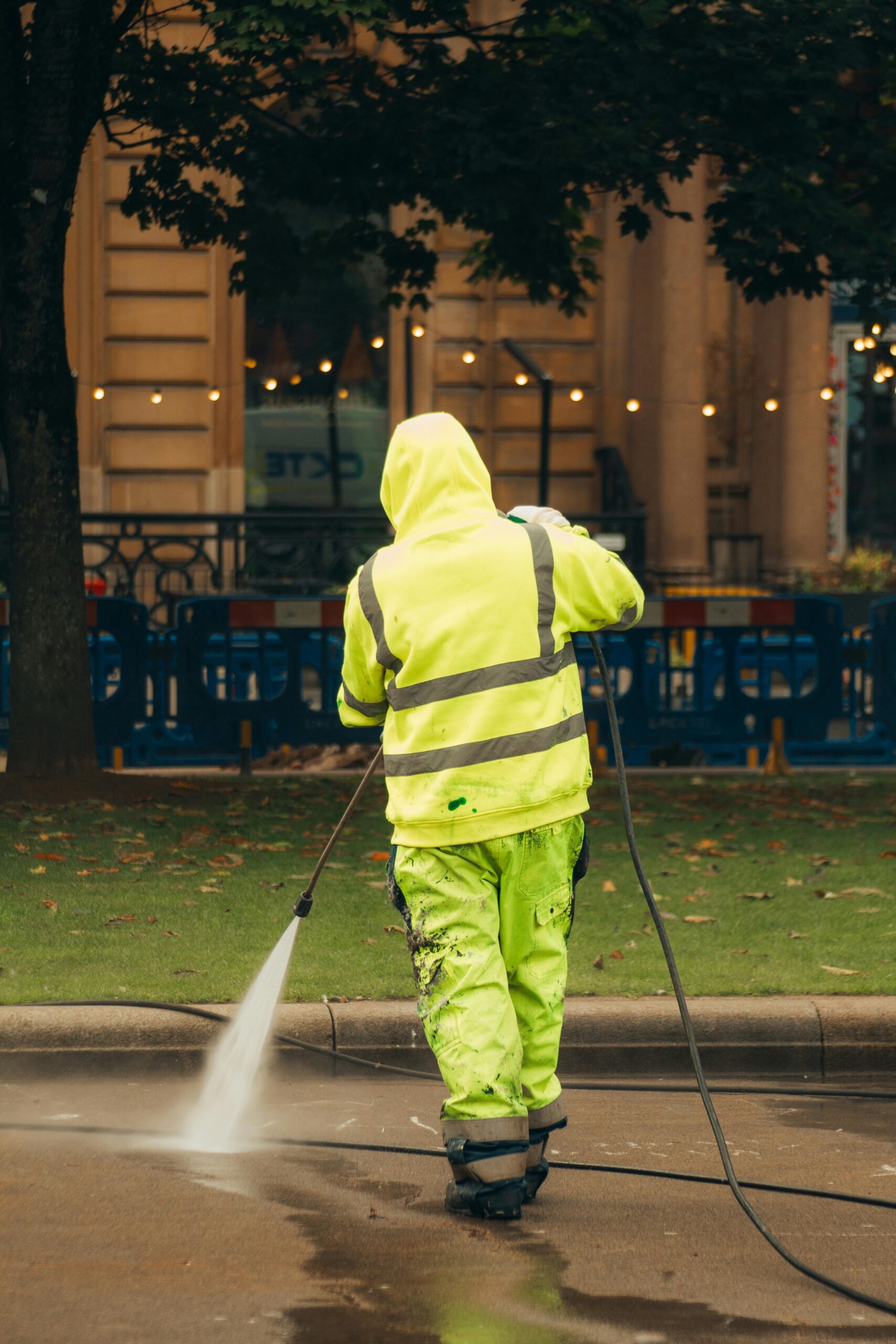 A city worker in high-visibility gear pressure washing a street corner.